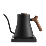 Matte black electric kettle with a wooden handle and lid knob, featuring a slender gooseneck spout, sitting on a square.
