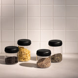 Four clear cylindrical vacuum canisters with black lids holding coffee beans, pasta, grains, and dried herbs on a tiled.