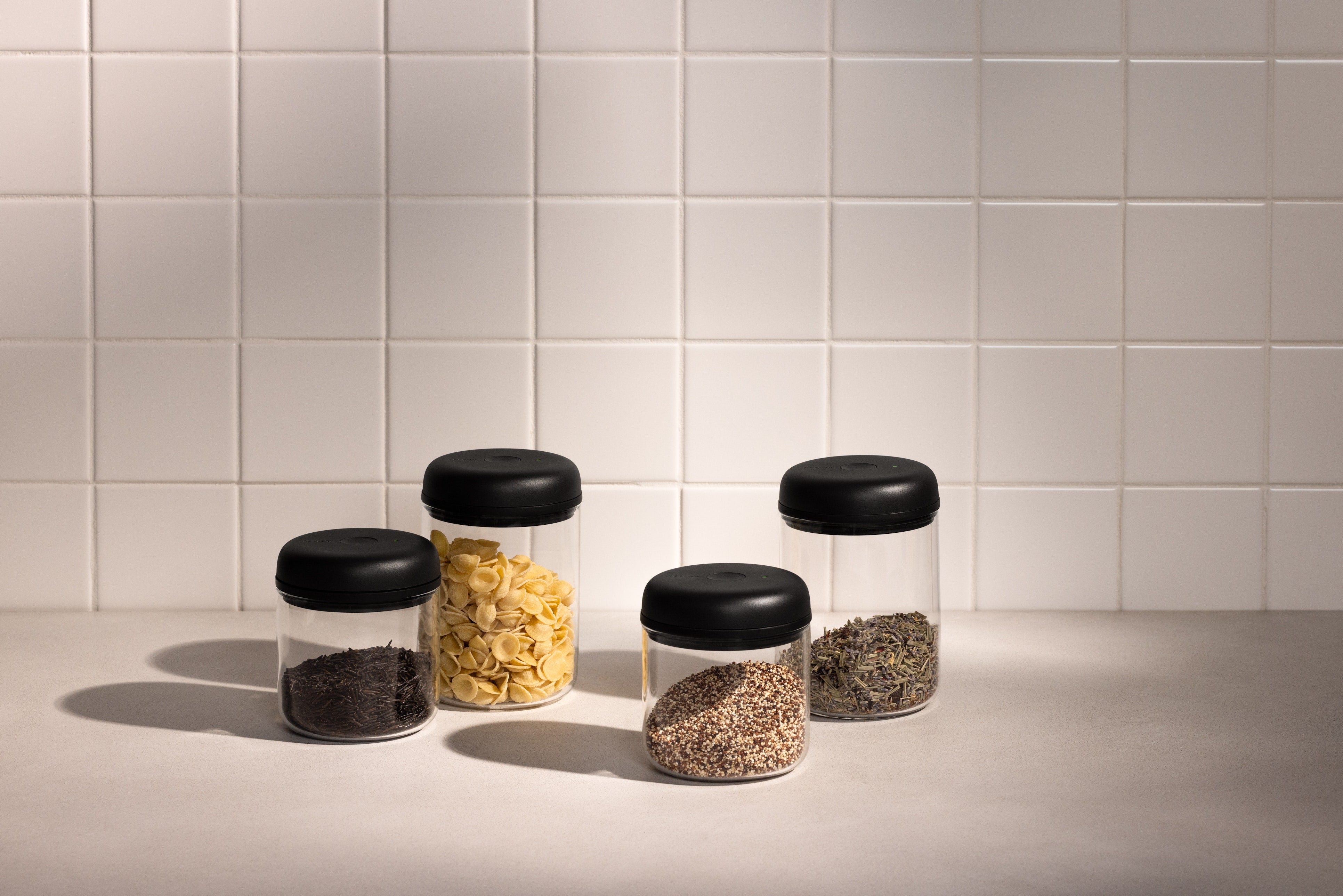 Four clear cylindrical vacuum canisters with black lids holding coffee beans, pasta, grains, and dried herbs on a tiled.
