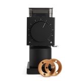 Matte black coffee grinder with large dial, transparent hopper, and two wooden burrs displayed in front.