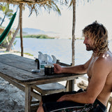 Man with tousled blonde hair sitting shirtless at weathered wooden table on sandy beach, holding black coffee mug next to.