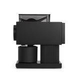 Matte black compact coffee grinder with transparent hopper, cylindrical stainless steel body, and ribbed grip base.