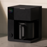 Compact matte black precision coffee maker with a cylindrical carafe and minimalist control knob.