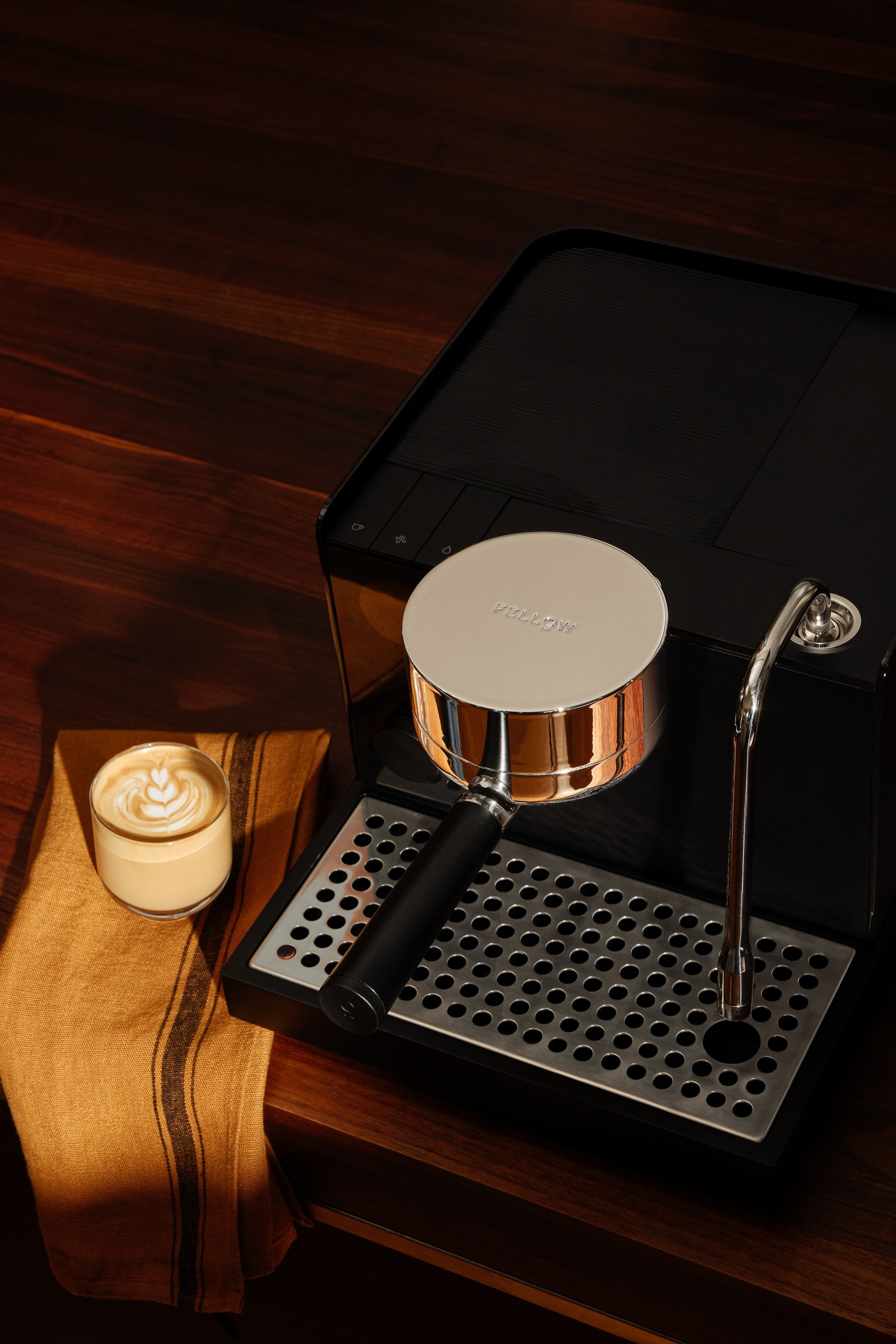 Matte black espresso machine with silver accents, steam wand, and drip tray on wooden surface with latte and cloth nearby.