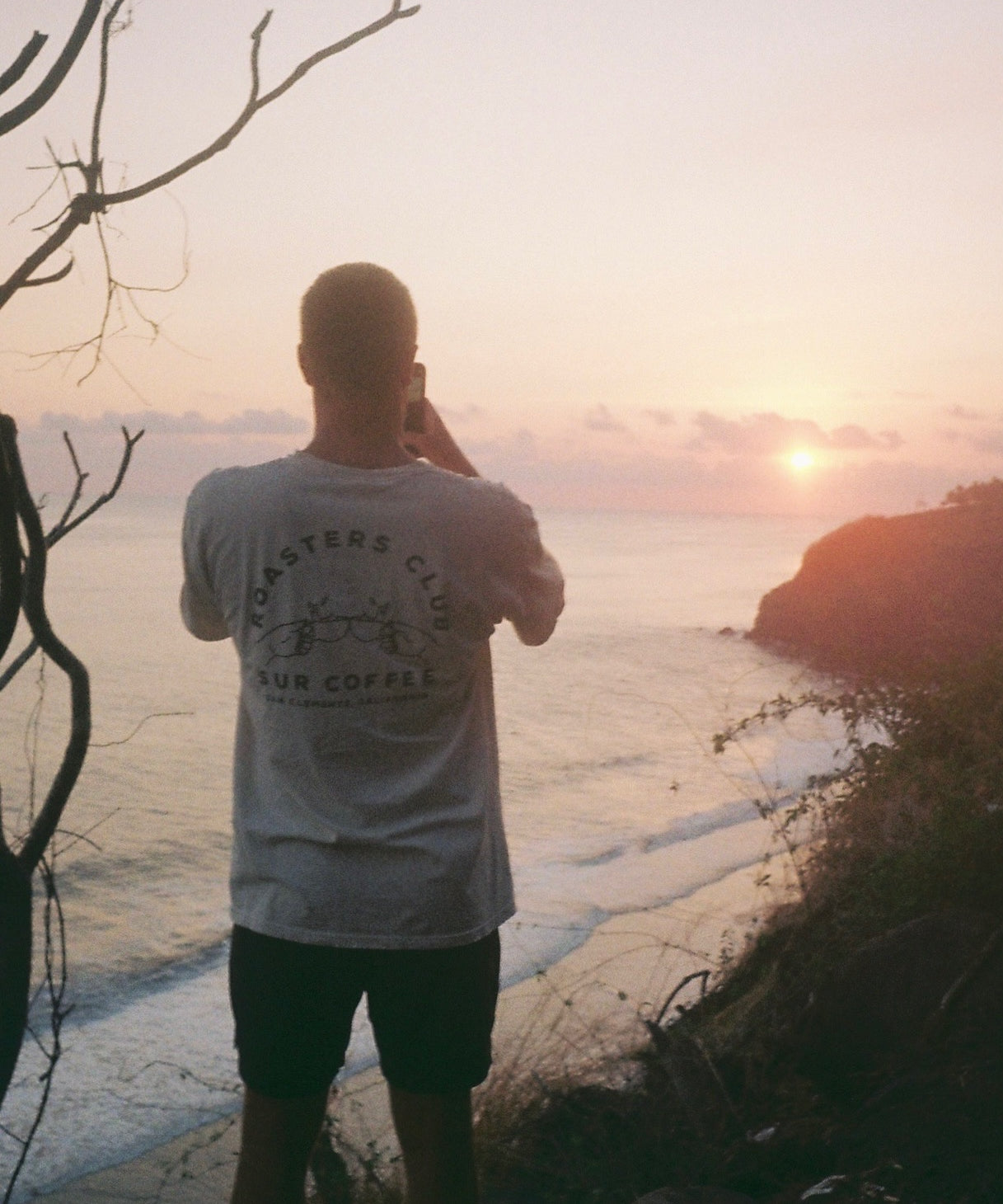 Light gray Roasters Club T-shirt with black graphic print on the back, worn by a person overlooking a coastal sunset.