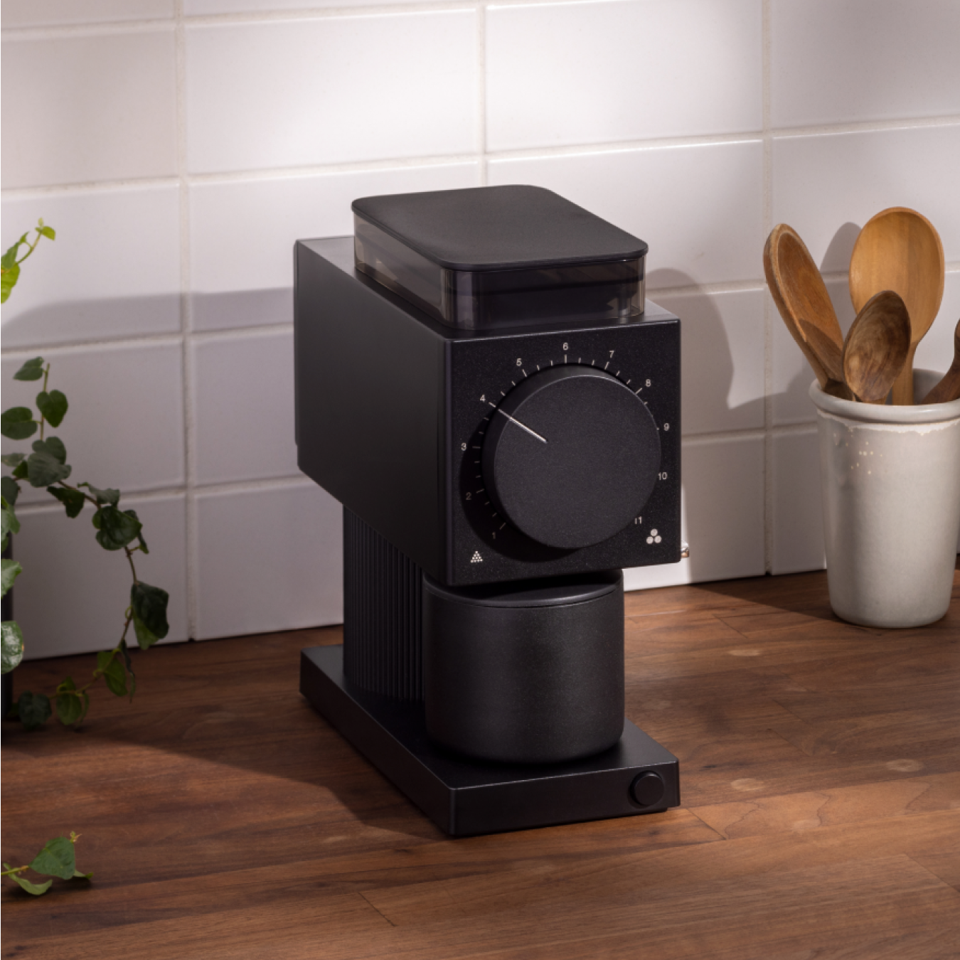 Matte black rectangular coffee grinder with large front dial and cylindrical grounds container on wooden counter.