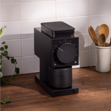 Matte black rectangular coffee grinder with large front dial and cylindrical grounds container on wooden counter.
