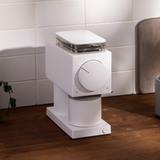 Compact white coffee grinder with clear lid, large front dial, and cylindrical base on wooden countertop.