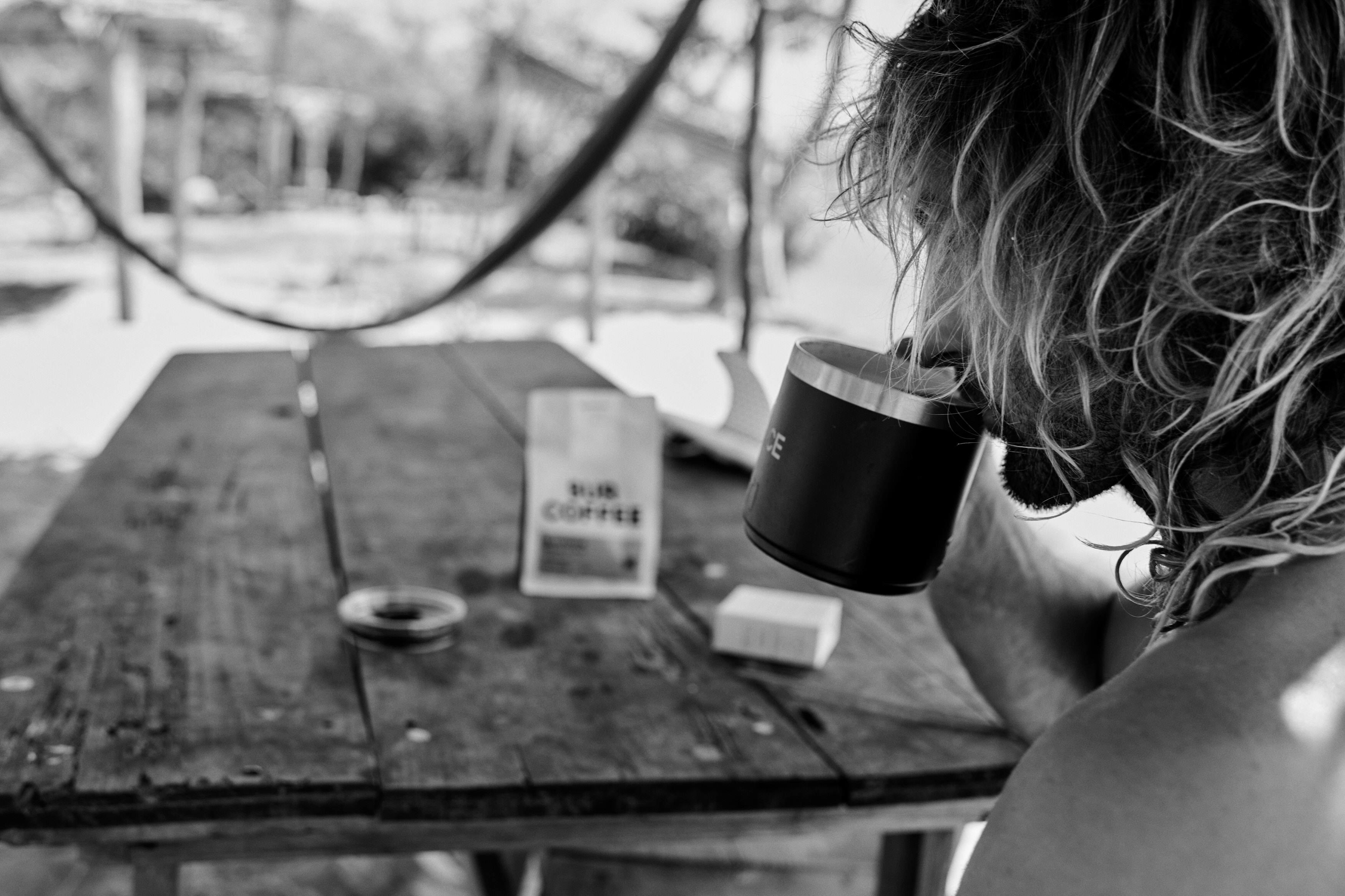 Black and white photo of a man with curly hair drinking coffee at a rustic wooden table outdoors.