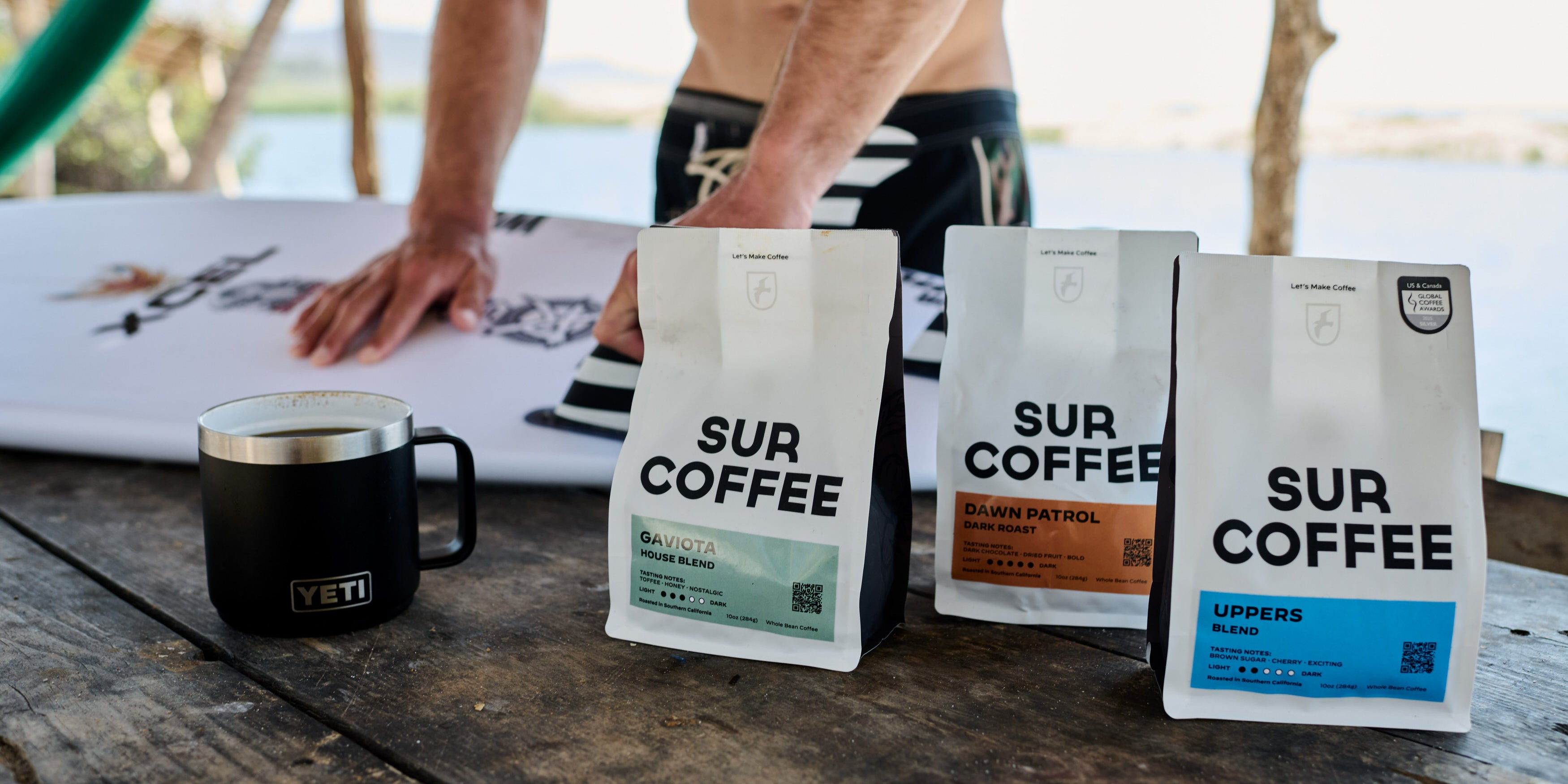 Three white bags of Sur Coffee blends on a wooden table with a black Yeti mug and a surfer.