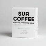 A white box of SUR COFFEE 'Ritual of Adventure Blend' specialty instant coffee, standing on a weathered wooden surface with a blurred outdoor background of green foliage.