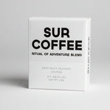 A white box of SUR COFFEE 'Ritual of Adventure Blend' specialty instant coffee, standing on a weathered wooden surface with a blurred outdoor background of green foliage.