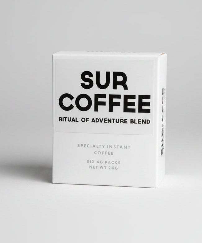 A white box of SUR COFFEE 'Ritual of Adventure Blend' specialty instant coffee, standing on a weathered wooden surface with a blurred outdoor background of green foliage.