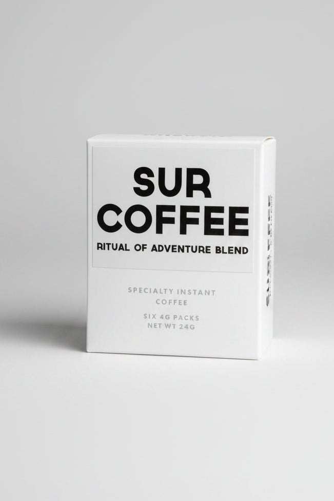 A white box of SUR COFFEE 'Ritual of Adventure Blend' specialty instant coffee, standing on a weathered wooden surface with a blurred outdoor background of green foliage.