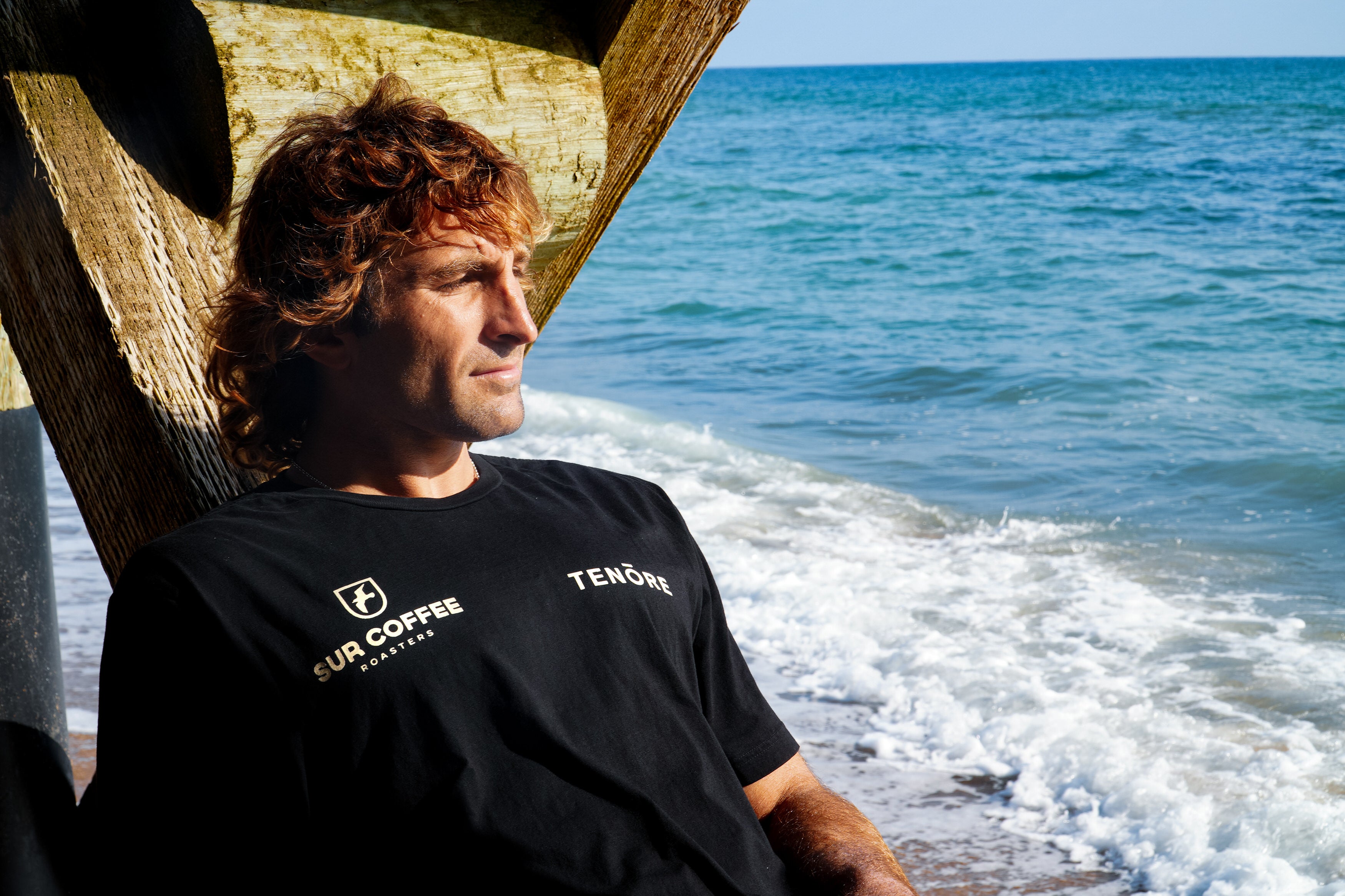Black Sur Coffee x Tenore tee with white logo and text, worn by a man sitting under a wooden structure by the ocean.
