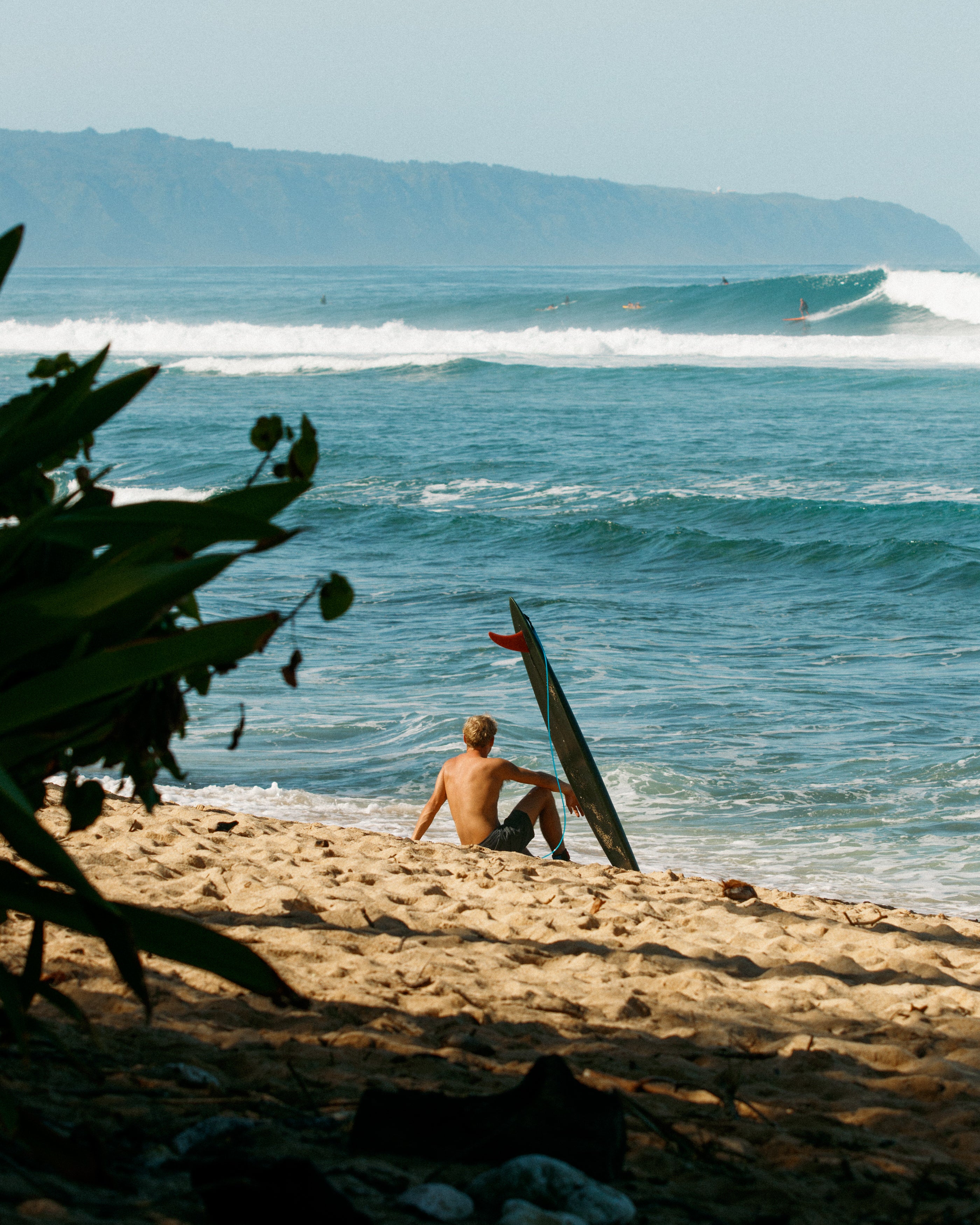 Person sitting on a sandy beach with a surfboard, looking at the ocean waves.